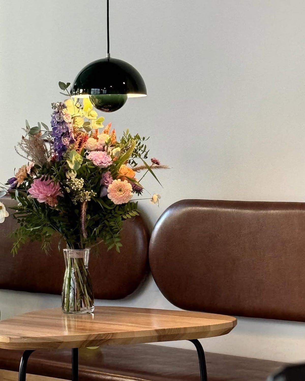 Stylowe wnętrze kawiarni z eleganckim, minimalistycznym wystrojem. W centrum kadru znajduje się bukiet świeżych kwiatów w szklanym wazonie – kolorowe dalie, róże i inne sezonowe rośliny dodają przestrzeni przytulnego charakteru. Bukiet stoi na drewnianym stoliku o organicznym kształcie, który komponuje się z brązową skórzaną ławką z owalnymi oparciami w tle. Nad stołem zawieszona jest ciemnozielona, metaliczna lampa w kształcie półkuli, emitująca ciepłe światło, podkreślające naturalne barwy wnętrza.