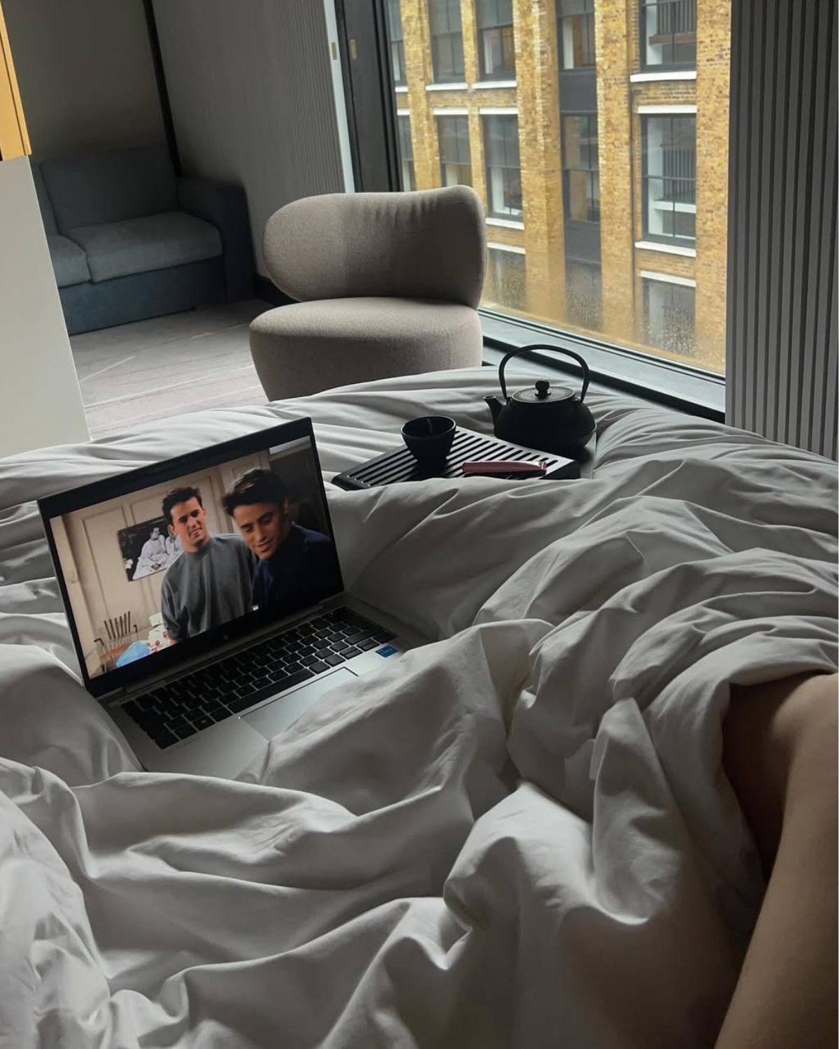 Zdjęcie przedstawiające łóżko z laptopem, na którym kobieta ogląda serial. Wykorzystała kody na Netflix 2025, by zobaczyć więcej tytułów. W tle widać tacę z dzbankiem i filiżanką herbaty, a także okno z widokiem na sąsiedni budynek.