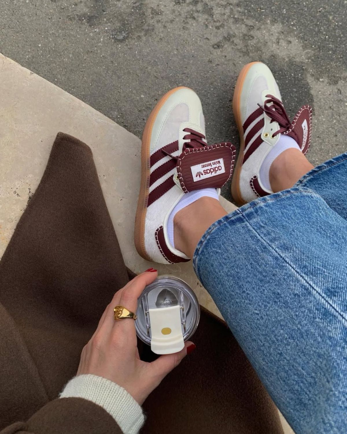 Zdjęcie przedstawia kolorowe sneakersy na wiosnę, czyli adidas samba w kolorze biało-bordowym. Zdjęcie robione jest z góry i jest skoncentrowane na obuwie. Widać fragment jeansów oraz brązowego płaszcza, a także rękę trzymającą kubek z kawą.