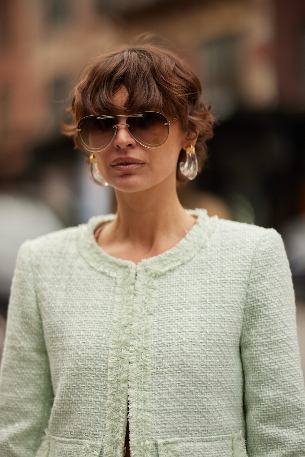 Elegancki street style – kobieta w tweedowej marynarce w pastelowym odcieniu zieleni– Kobieta o krótkiej fryzurze i pełnych, falujących grzywkach pozuje na tle miejskiej ulicy. Ma na sobie elegancką, jasnozieloną marynarkę wykonaną z tweedu, którą łączy z dużymi, geometrycznymi kolczykami i stylowymi okularami przeciwsłonecznymi z metalowymi oprawkami. Kolory na wiosnę 2025 zachwycają w jej wykonaniu.