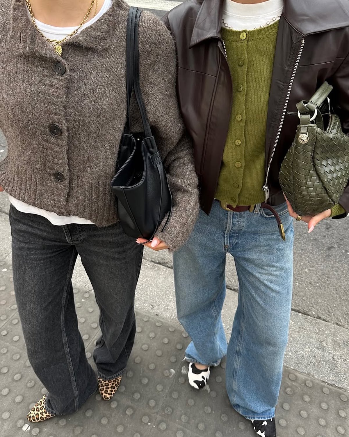 Dwie osoby trzymające się za ręce, ubrane w stylizacje łączące elementy retro i nowoczesnej elegancji. Osoba po lewej ma na sobie melanżowy, wełniany kardigan w odcieniu taupe z dużymi guzikami, biały t-shirt pod spodem, czarne dżinsy oraz buty z czubkiem w panterkowy wzór. Stylizację uzupełnia złoty naszyjnik z liściem i czarna, skórzana torebka o minimalistycznym fasonie.