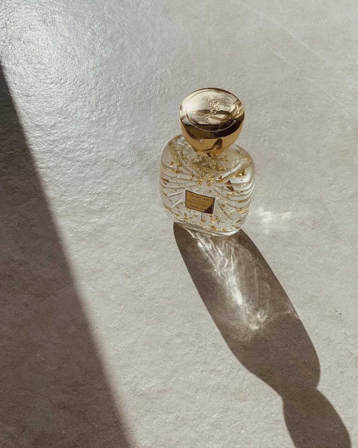 Perfumy, które pachną na każdym inaczej – na zdjęciu widać flakon perfum w przezroczystym flakonie ze złotą zatyczką. Stoją na betonowym blacie i oświetla je blask słońca.