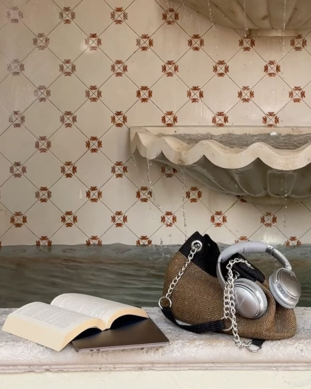 Polskie podcasty o modzie: zdjęcie przedstawiające brzeg fontanny, na którym leży torebka, słuchawki bezprzewodowe, a także książka i laptop.