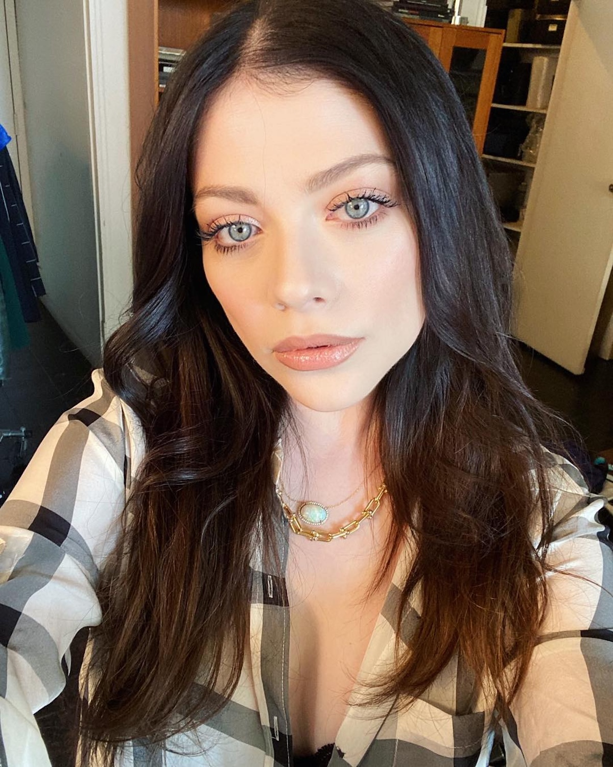 Michelle Trachtenberg przyczyna śmierci: Zbliżenie na młodą kobietę o jasnej cerze, długich falujących ciemnych włosach i wyrazistych niebieskich oczach. Ma delikatny makijaż podkreślający rzęsy oraz subtelnie połyskujące usta w odcieniu nude. Nosi koszulę w czarno-białą kratę oraz złoty naszyjnik z opalizującym kamieniem. W tle widać otwartą szafę oraz półki z książkami, co sugeruje domowe otoczenie.