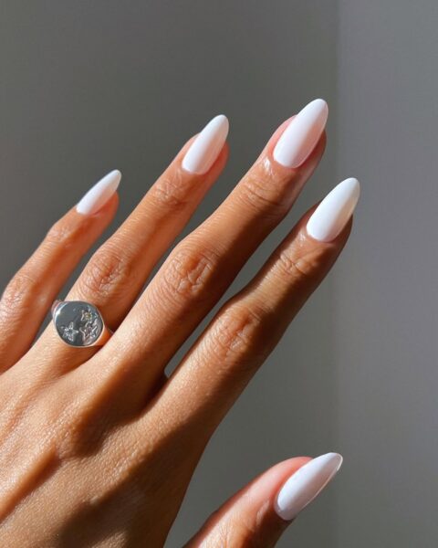 Minimalistyczny biały manicure na długich paznokciach – Bliskie ujęcie dłoni z perfekcyjnie pomalowanymi paznokciami w odcieniu czystej bieli. Smukłe, migdałowe kształty paznokci pięknie komponują się z subtelnym srebrnym pierścionkiem z gwiazdami.