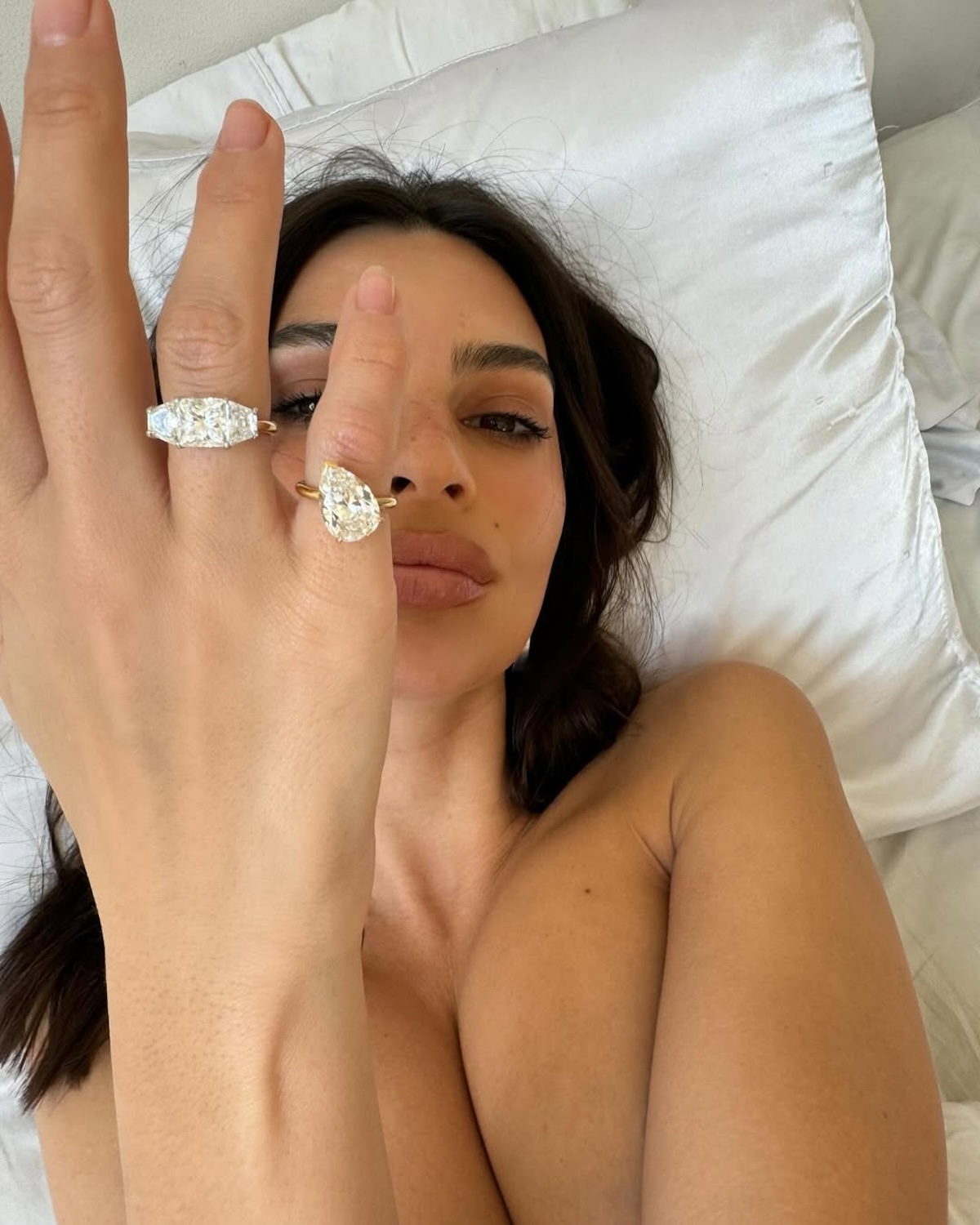Emily Ratajkowski leżąca na łóżku prezentuje dwa duże pierścionki zaręczynowe na palcach – jeden w kształcie gruszki, drugi o klasycznym, prostokątnym szlifie. Idealny wybór pierścionka zaręczynowego powinien uwzględniać styl i preferencje przyszłej narzeczonej. Jak wybrać pierścionek zaręczynowy, który podkreśli osobowość ukochanej?