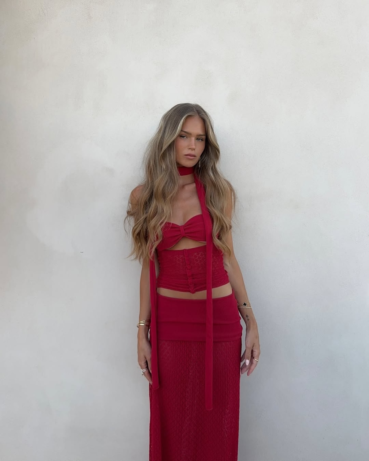 Stylizacja składająca się z czerwonego crop topu z wiązaniem i długiej, koronkowej spódnicy. Modelka w letniej stylizacji na tle jasnej ściany, podkreślającej intensywny odcień czerwieni.