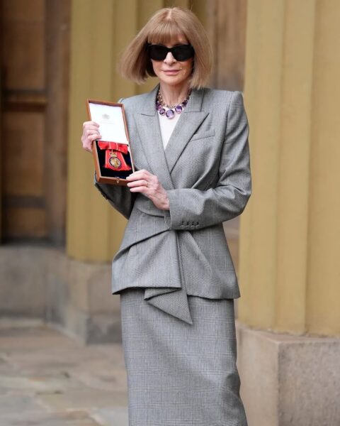 Anna Wintour 2025: Anna Wintour odbierająca nagrodę Order Companions of Honour w eleganckim szarym garniturze od Alexandra McQueena z kratką stoi przed historycznym budynkiem, trzymając otwarte pudełko z odznaczeniem Orderu Imperium Brytyjskiego. Ma na sobie ciemne okulary przeciwsłoneczne oraz fioletowy naszyjnik, podkreślający jej stylowy wygląd.
