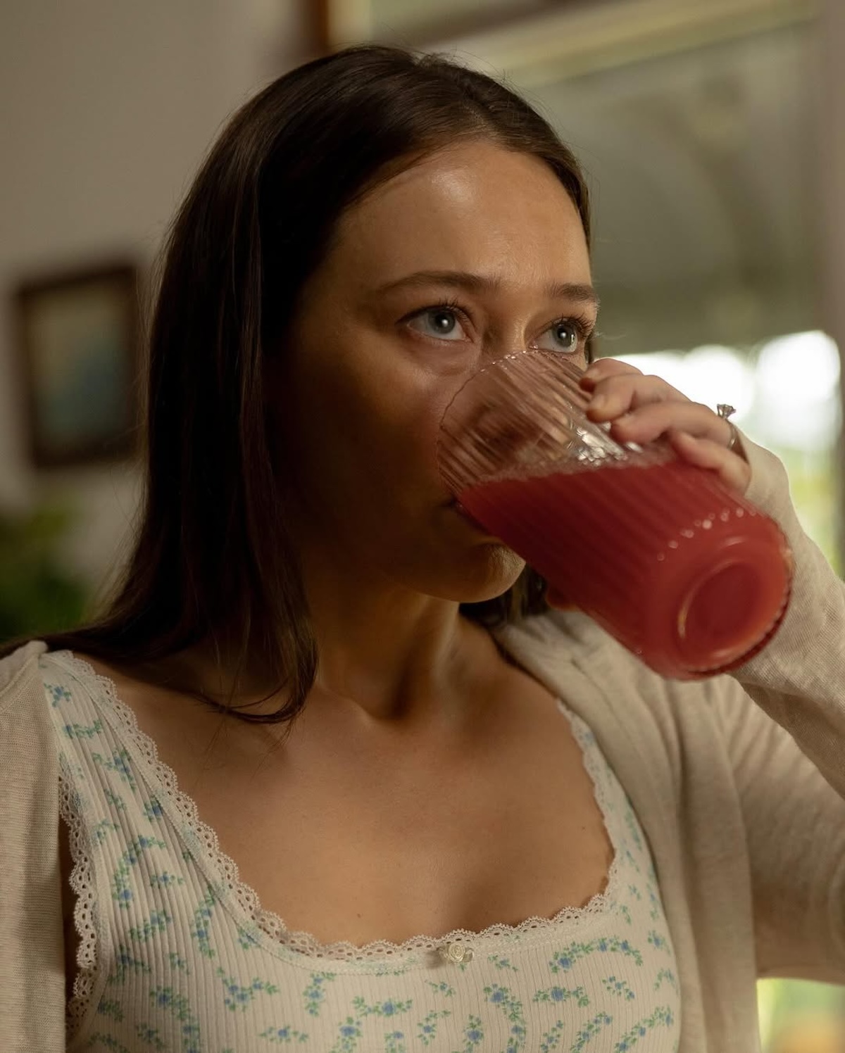 Kadr z serialu „Apple Cider Vinegar” – tajemniczy moment – Bohaterka grana przez Alycię Debnam-Carey pije czerwony napój z przezroczystej szklanki. Jej intensywne spojrzenie sugeruje, że scena jest kluczowa dla fabuły serialu. Subtelna stylizacja w pastelowych kolorach i romantyczna sceneria w tle dodają jej delikatności i tajemniczości. Milla Blake kto to