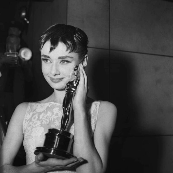 Audrey Hepburn ze statuetką Oscara po ceremonii wręczenia nagród w 1954 roku. Aktorka, ubrana w elegancką białą suknię z koronkowymi detalami od Givenchy, trzyma nagrodę za najlepszą rolę pierwszoplanową w filmie Rzymskie wakacje. Jej krótkie włosy są ułożone w charakterystyczny pixie cut, a delikatny uśmiech i wzruszone spojrzenie podkreślają emocjonalny moment. W tle fotografowie uchwytują tę historyczną chwilę.