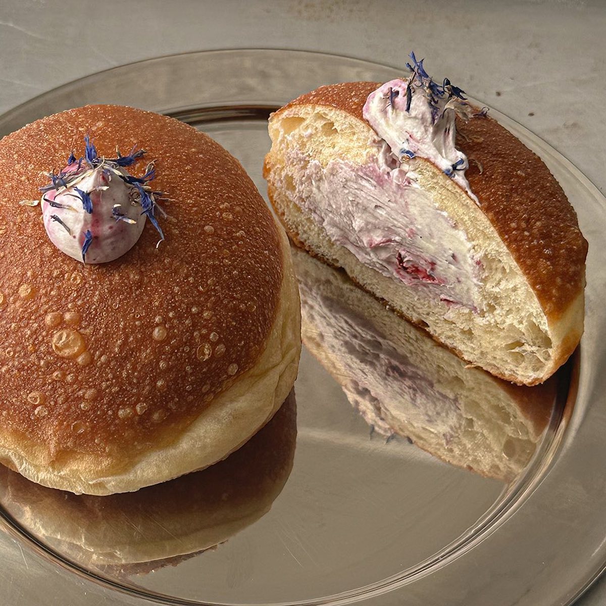 Na zdjęciu widoczne są dwa wegańskie pączki na metalowej tacy. Jeden pączek jest cały, ozdobiony kremem i płatkami kwiatów, a drugi przekrojony, ukazując puszyste nadzienie o jasnoróżowym odcieniu z kawałkami owoców. Idealnie miękkie ciasto i artystyczne zdobienie podkreślają wyjątkowy charakter tego deseru.