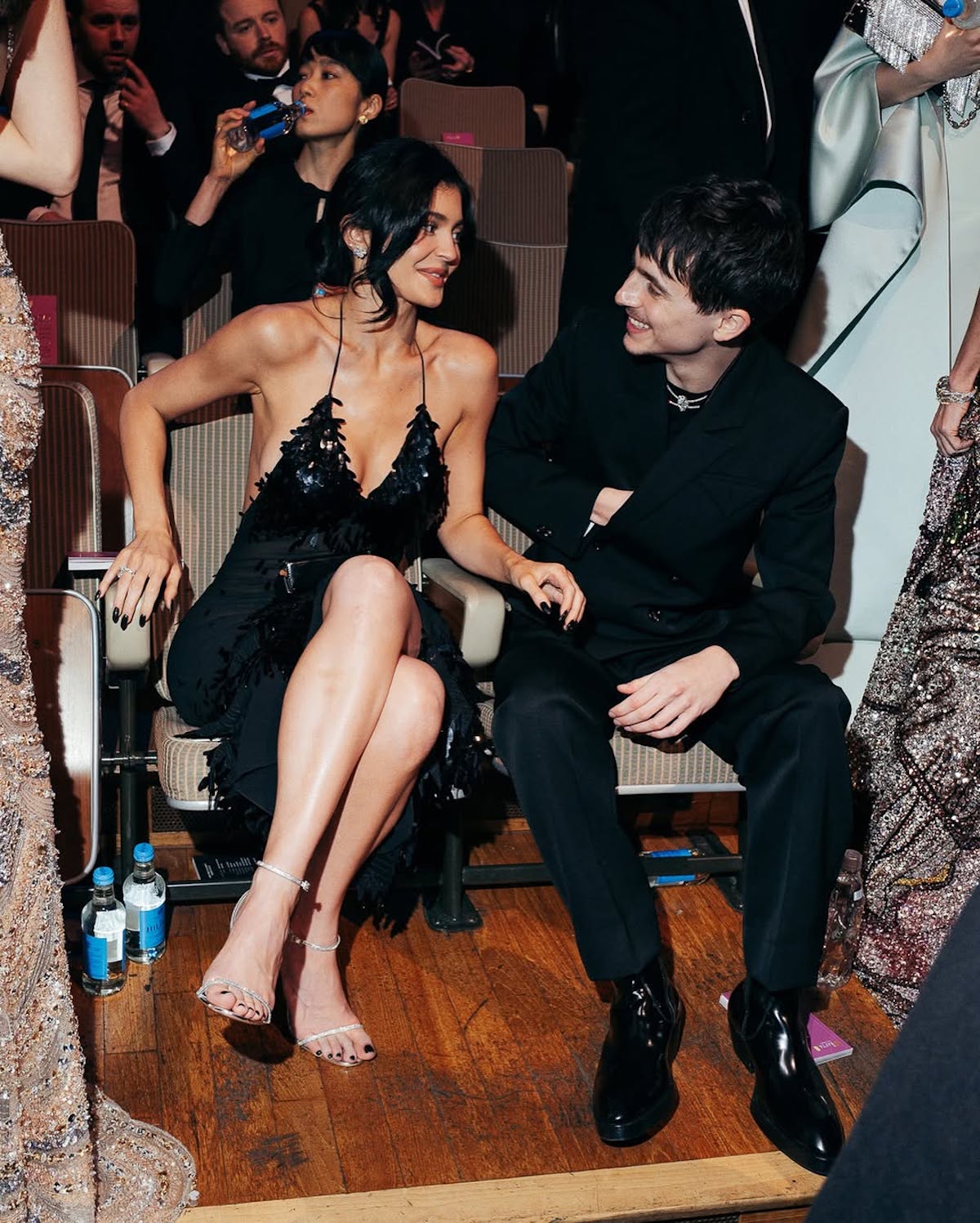 Kylie Jenner i Timothée Chalamet siedzą razem na eleganckim wydarzeniu, uśmiechając się do siebie. Kylie ma na sobie czarną, błyszczącą sukienkę na ramiączkach z frędzlami, odsłaniającą ramiona i nogi, oraz srebrne sandałki na obcasie. Timothée jest ubrany w klasyczny, czarny garnitur i czarne, lakierowane buty. Oboje wyglądają na zrelaksowanych i zadowolonych, a wokół nich widać innych elegancko ubranych gości. Zdjęcie zostało zrobione podczas gali BAFTA 2025 w Londynie, dzień po ich wspólnym pojawieniu się na festiwalu Berlinale w Niemczech