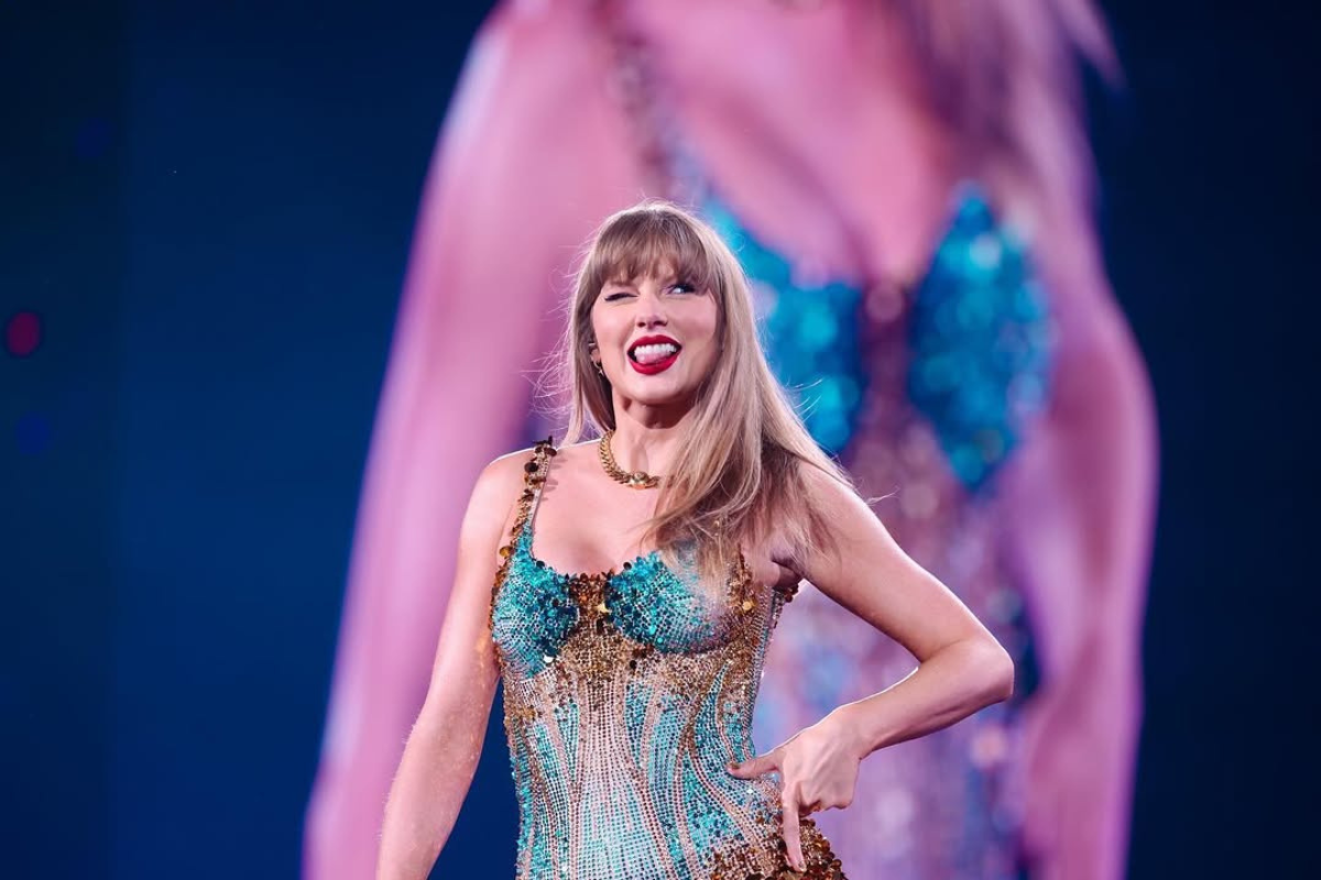 Taylor Swift w błyszczącej, zdobionej złotem i turkusowymi elementami sukni na scenie, uśmiecha się i puszcza oko w stronę publiczności. W tle widać powiększone jej odbicie na ekranie, co podkreśla efektowność występu.