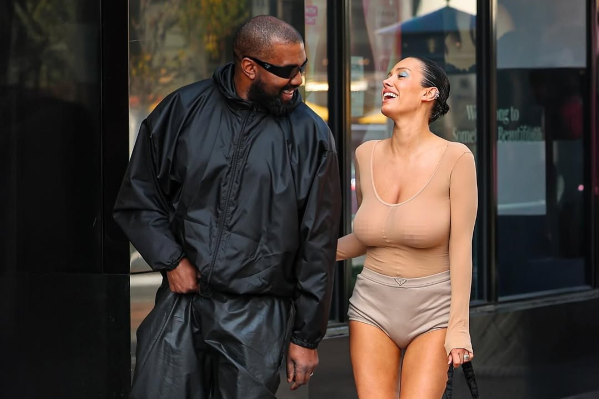 Bianca Censori bez cenzury uśmiechnięta podczas rozmowy z Kanye Westem – w cielistej, dopasowanej stylizacji z krótkimi szortami i białymi szpilkami, trzymająca czarną torebkę, u boku Kanye Westa w czarnym, sportowym zestawie.