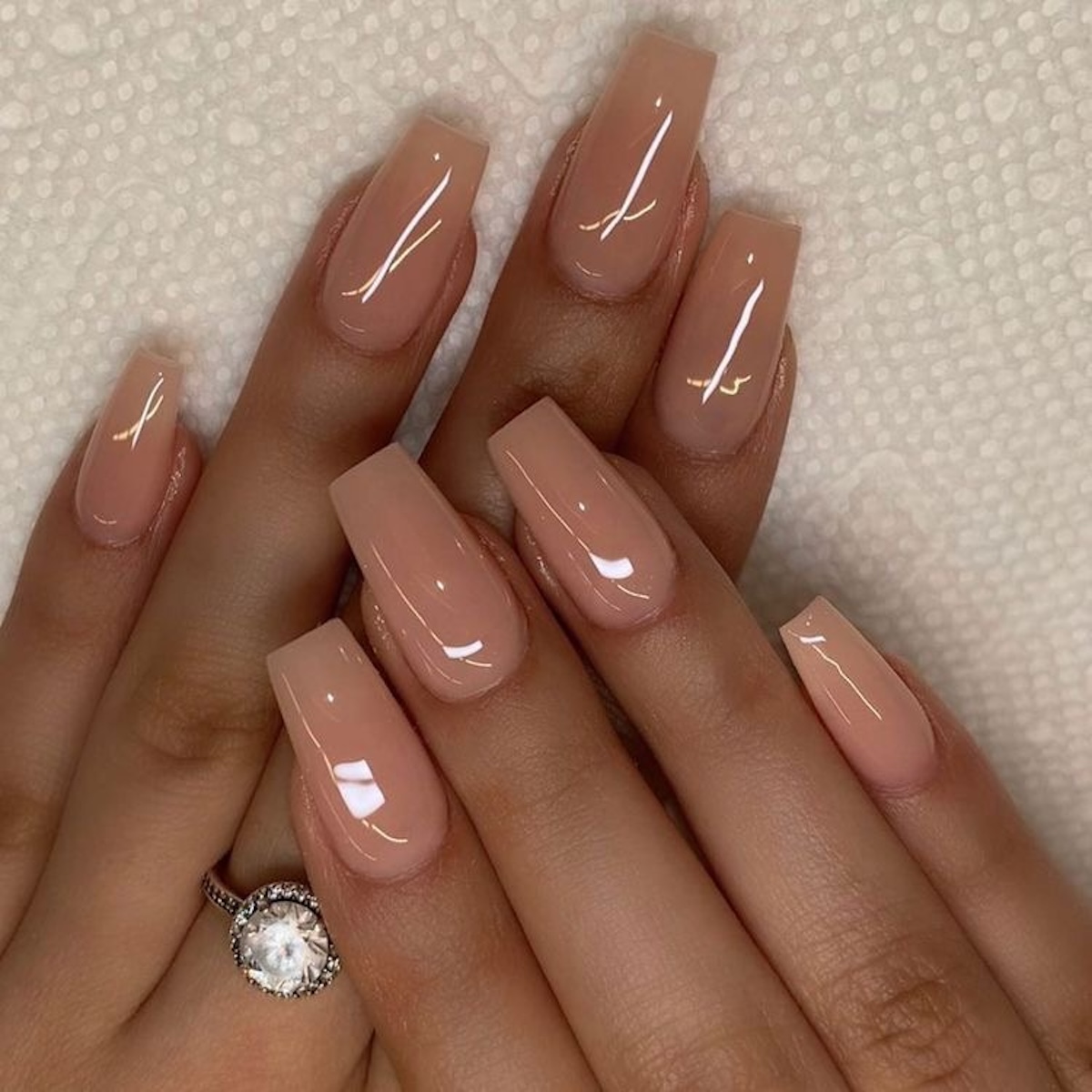 Paznokcie 2025: Dłonie z pięknie wymodelowanymi, kwadratowymi paznokciami w odcieniu nude o błyszczącym wykończeniu. Subtelny i elegancki manicure nadaje dłoniom zadbany i klasyczny wygląd. Na palcu znajduje się srebrny pierścionek z dużym diamentem.