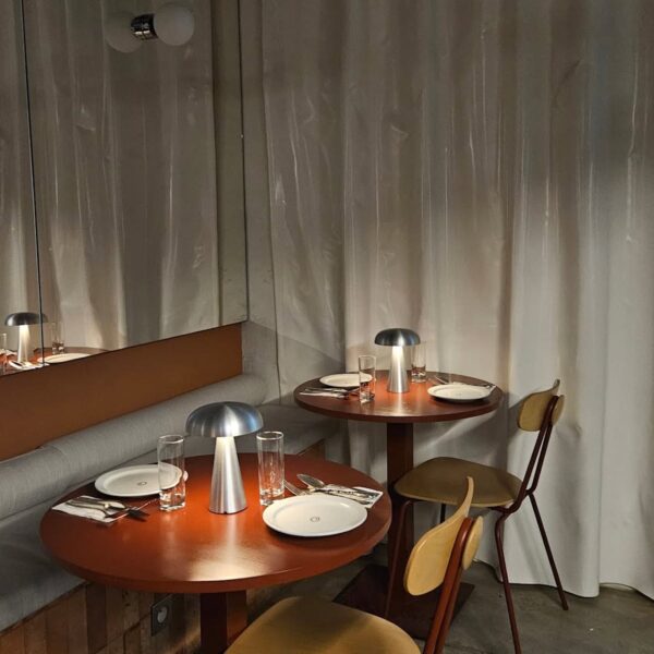 Dwa elegancko nakryte, okrągłe stoliki w restauracji, ustawione przy ścianie z zasłoną i dużym lustrem. Każdy stolik ma dwie drewniane krzesła z metalowymi nogami, proste nakrycie z talerzami, sztućcami i szklankami oraz nowoczesną, srebrną lampkę stołową o miękkim świetle. W tle widoczne tapicerowane siedzisko.