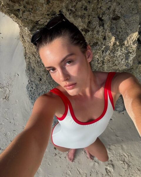 Kobieta w białym jednoczęściowym stroju kąpielowym z czerwonymi lamówkami robi selfie na plaży przy skalistej ścianie. Ma ciemne, zaczesane do tyłu włosy i okulary przeciwsłoneczne na głowie. Stoi boso na jasnym piasku w naturalnym świetle słonecznym.
