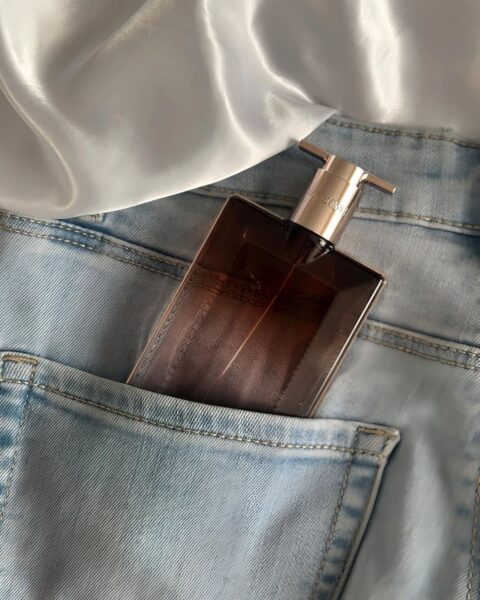 Perfumy na wiosnę 2025 od Lancôme w kwadratowym flakonie ze złotą zatyczką. Flakon schowany jest w tylną kieszeń jasnych, błękitnych jeansów. Widać fragment białej koszuli wsuniętej w pas.