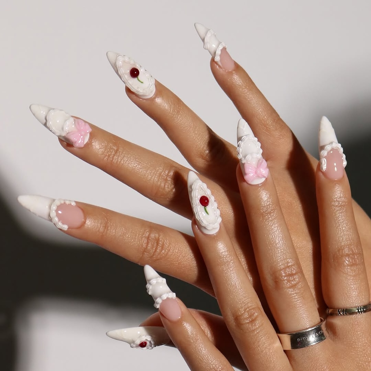 Lukrowane paznokcie: Dłonie z jasnoróżowym manicure o strukturalnym wykończeniu. Paznokcie są udekorowane białymi koronkowymi obramowaniami oraz miniaturowymi kokardkami i serduszkami. Stylizacja łączy estetykę retro z romantycznym, dziewczęcym akcentem.