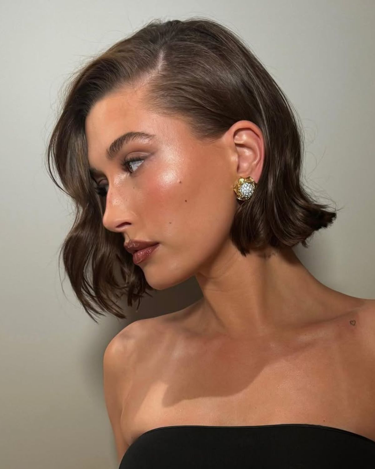 Zdjęcie Hailey Bieber, która prezentuje trendy w koloryzacji włosów w marcu 2025: ciemne, jednolite włosy ścięte na boba. Ma przedziałek z boku, a w uszach kolczyki. Ubrana jest w czarny top bez ramiączek. Pozuje z boku.