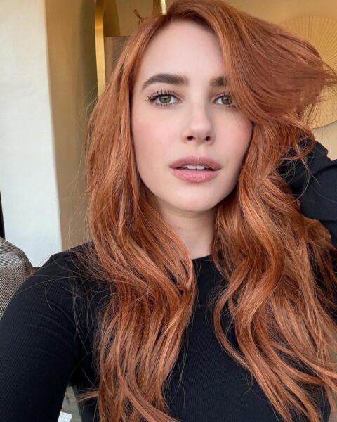 Aktorka Emma Roberts w kolorze terracopper, czyli ciepłym rudym odcieniu. Na zdjęciu ma rozpuszczone włosy. Ubrana jest w czarną bluzkę z długim rękawem. Za nią widać wnętrze mieszkania. Robi sobie selfie.