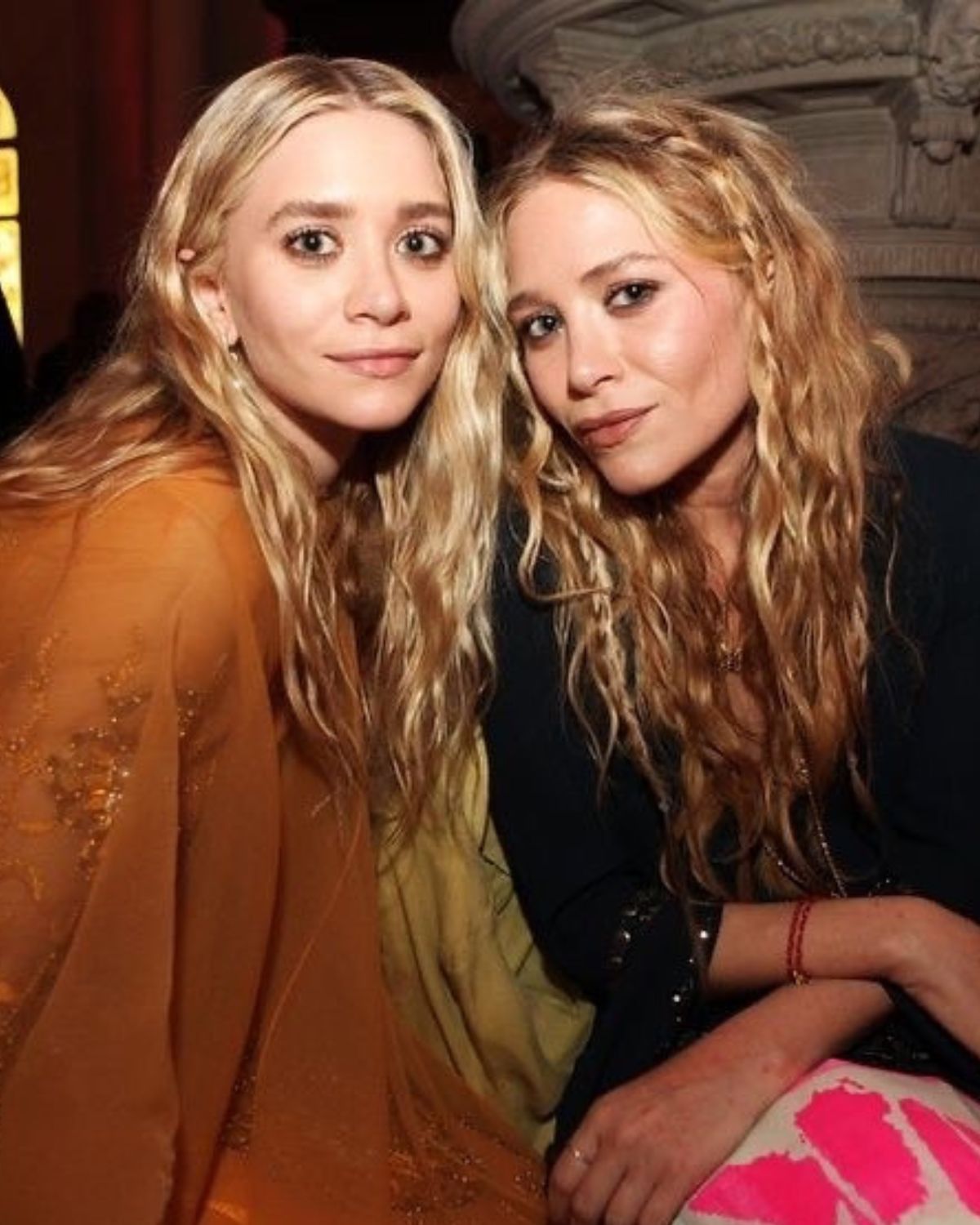 Zdjęcie sióstr Olsen, Mary-Kate i Ashley, które wylansowały włosy Olsen dip z dużym odrostem. Na zdjęciu siedzą obok siebie na evencie. Jedna ubrana jest w ceglaną sukienkę z tiulu, a druga ma na sobie czarną bluzkę oraz biało-różową spódnicę.