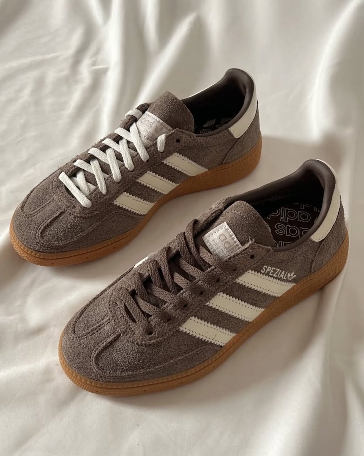 Para brązowych sneakersów Adidas Handball Spezial z białymi paskami i gumową podeszwą, leżących na jasnym materiale. Lewy but ma białe sznurówki, a prawy brązowe. Logo Adidas oraz napis 'Spezial' widoczne z boku butów.