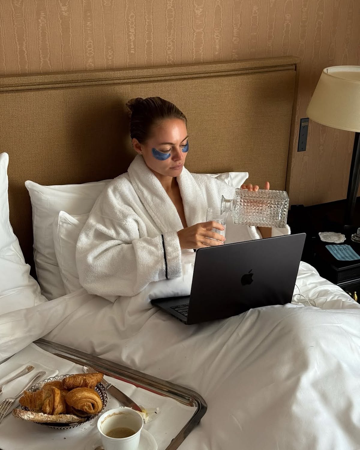 Kobieta w białym szlafroku siedzi w łóżku hotelowym z laptopem na kolanach, nosząc hydrożelowe płatki pod oczami. W jednej ręce trzyma szklankę, do której nalewa wodę z karafki. Przed nią na tacy znajduje się śniadanie z rogalikami i filiżanką kawy