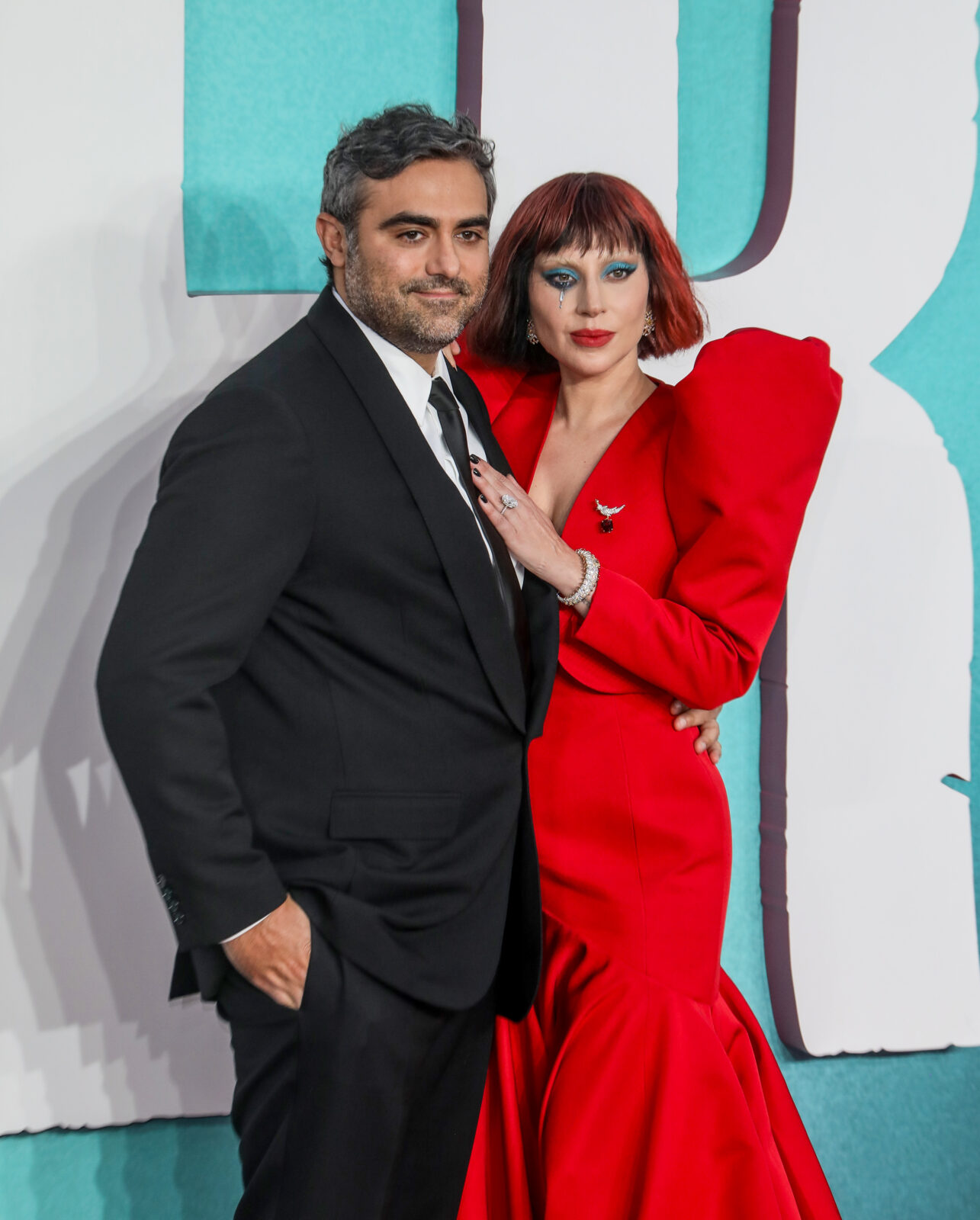Lady Gaga i Michael Polansky pozują razem na czerwonym dywanie podczas premiery. Gaga ubrana jest w spektakularną, czerwono-karminową suknię z dużymi ramionami i głębokim dekoltem. Ma krótkie, czerwone włosy i mocny makijaż z niebieskim cieniem i łzami z brokatu pod okiem. Polansky nosi klasyczny czarny garnitur z krawatem. Stoją blisko siebie, Gaga opiera dłoń na jego piersi.