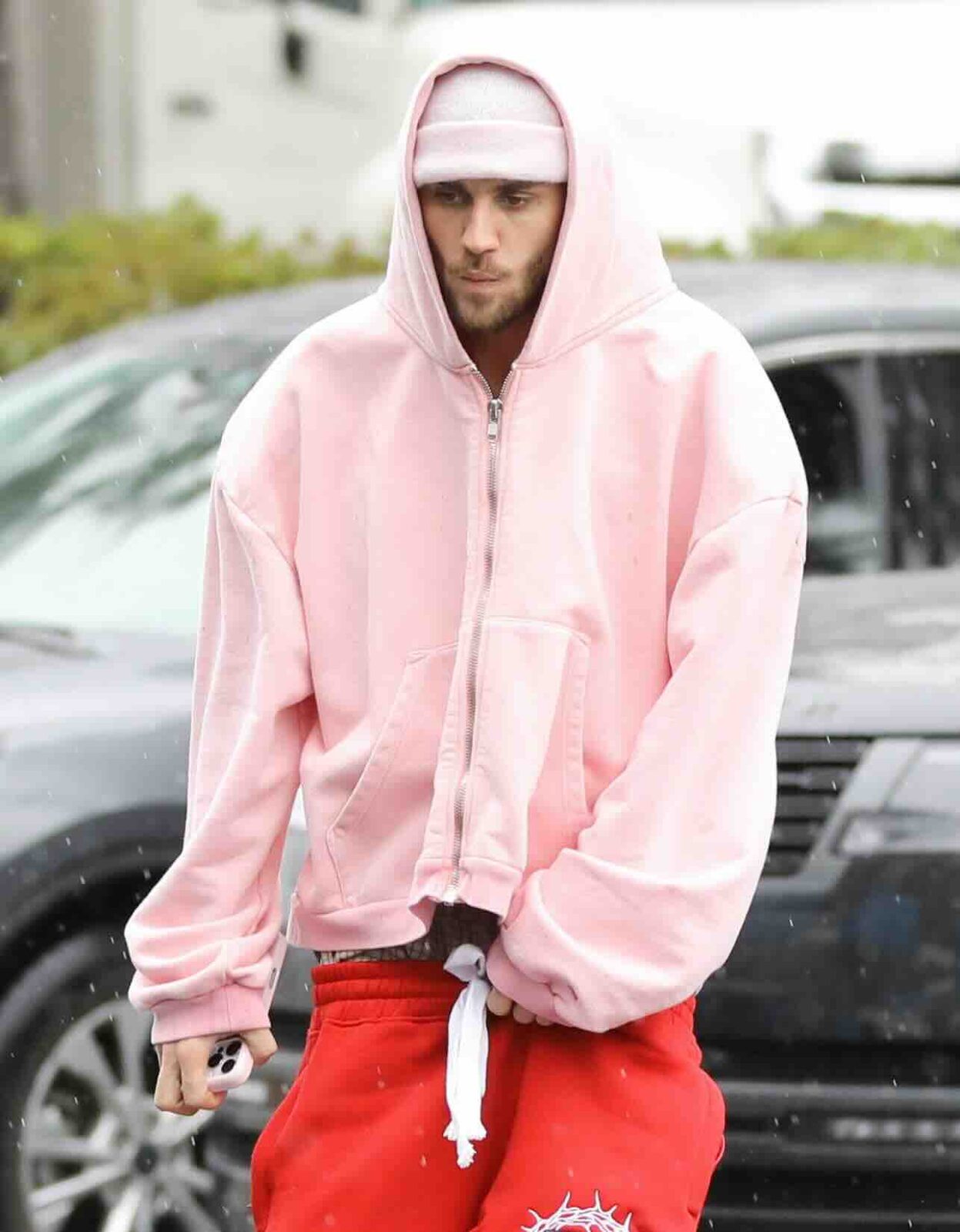 Justin Bieber w różowej bluzie z kapturem, czerwonych spodniach dresowych i opasce, uchwycony w deszczu – minimalistyczny, ale przemyślany look, balansujący między comfortwear a streetwearem nowej ery, której kuratorką jest Jenna Tyson.