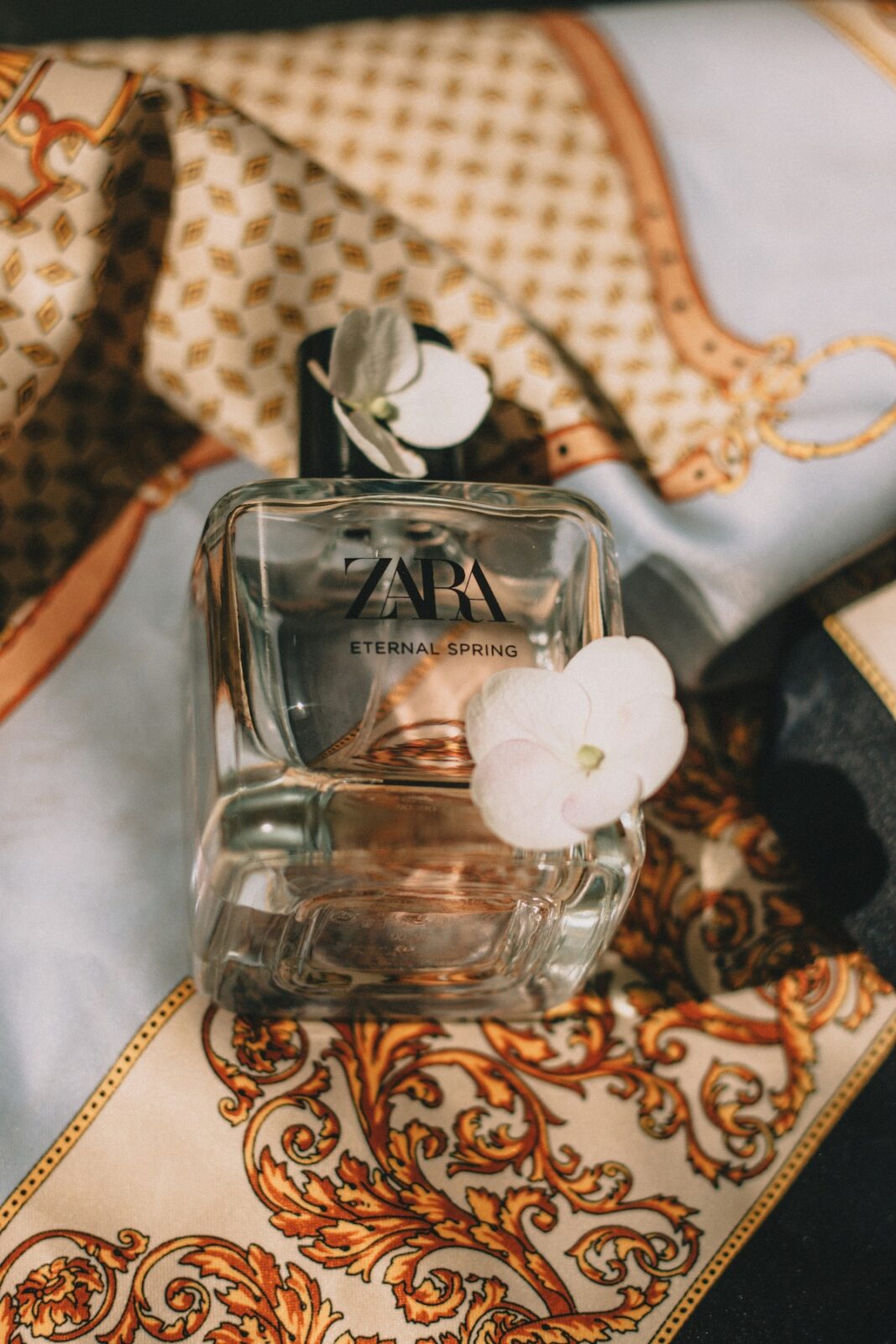 Perfumy z Zary: opinie. Na zdjęciu widać szklaną butelkę perfum "ZARA Eternal Spring", która leży na jedwabistej apaszce o eleganckim wzorze w odcieniach beżu, złota i błękitu. Na butelce leżą białe kwiaty, dodając kompozycji romantycznego akcentu. Światło pada na szkło i tkaninę.