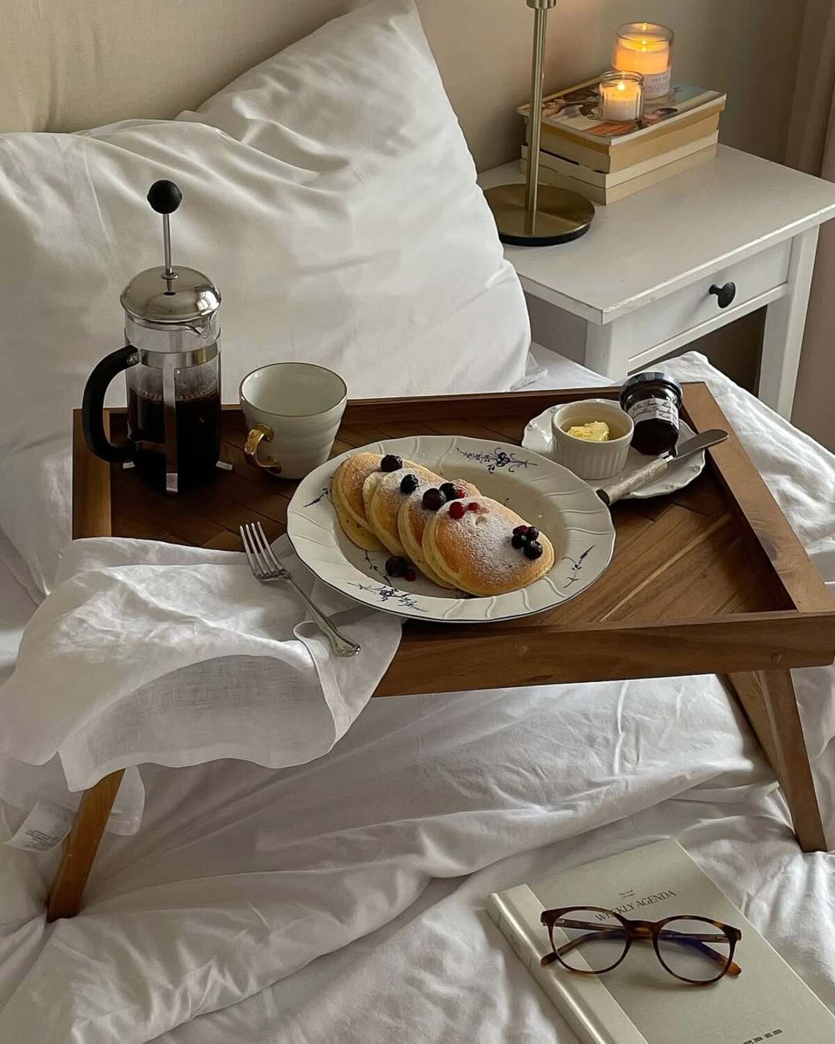 Drewniana taca na białej pościeli z french pressem, talerzem pancakes udekorowanych jagodami, małą filiżanką, kostką masła, świecą na stoliku nocnym oraz okularami leżącymi na książce. Zdjęcie przedstawia śniadaniowe grzechy, których należy unikać.