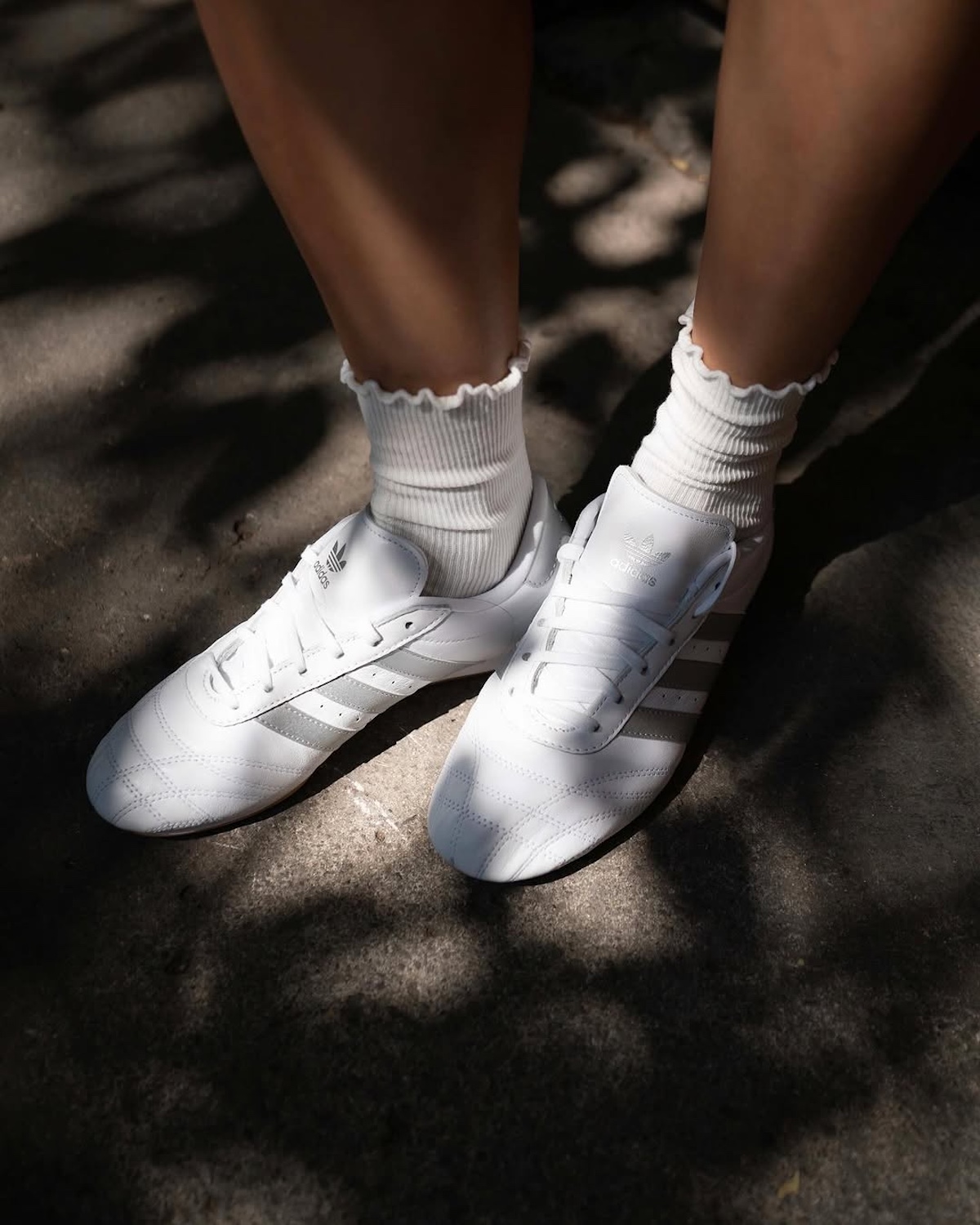 Zbliżenie na zdjęcie jednych z najmodniejszych adidasów na wiosnę 2025. To model adidas Taekwondo w kolorze białym, które modelka połączyła z białymi skarpetkami z falbankami. Na buty i ulicę pada cień.