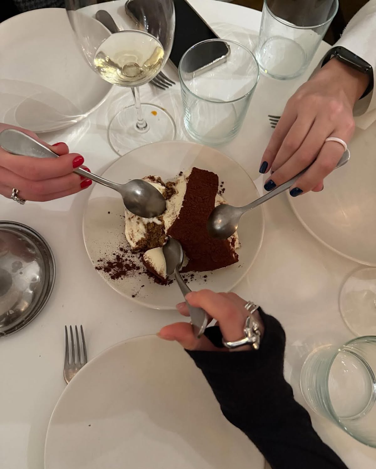Na zdjęciu widać trzy osoby dzielące się deserem tiramisu, każda z nich trzyma łyżeczkę i sięga po porcję ze wspólnego talerza. Dłonie ozdobione są różnymi pierścionkami i mają pomalowane paznokcie w różnych kolorach (czerwony, granatowy, naturalny). Na stole znajduje się kieliszek z białym winem, kilka pustych szklanek i talerzy oraz telefon komórkowy. Całość utrzymana jest w ciepłej, towarzyskiej atmosferze przy wspólnym stole.