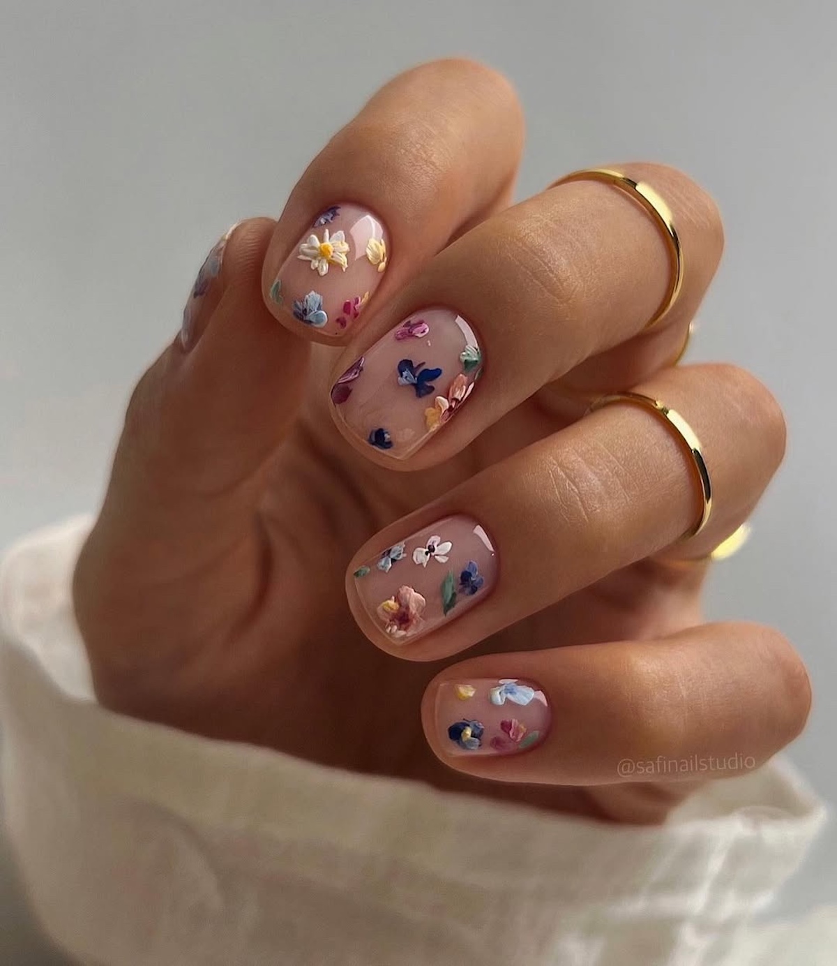 Paznokcie na Wielkanoc wzory w kwiaty 2025. Zbliżenie na dłoń z transparentnym manicurem, na którym namalowane są małe, kolorowe kwiatki. Paznokcie są owalne, a na palcach znajdują się dwa złote pierścionki. W prawym dolnym rogu widoczny jest napis "@safinallstudio". Dłoń jest oświetlona jasnym światłem, a tło jest szare.