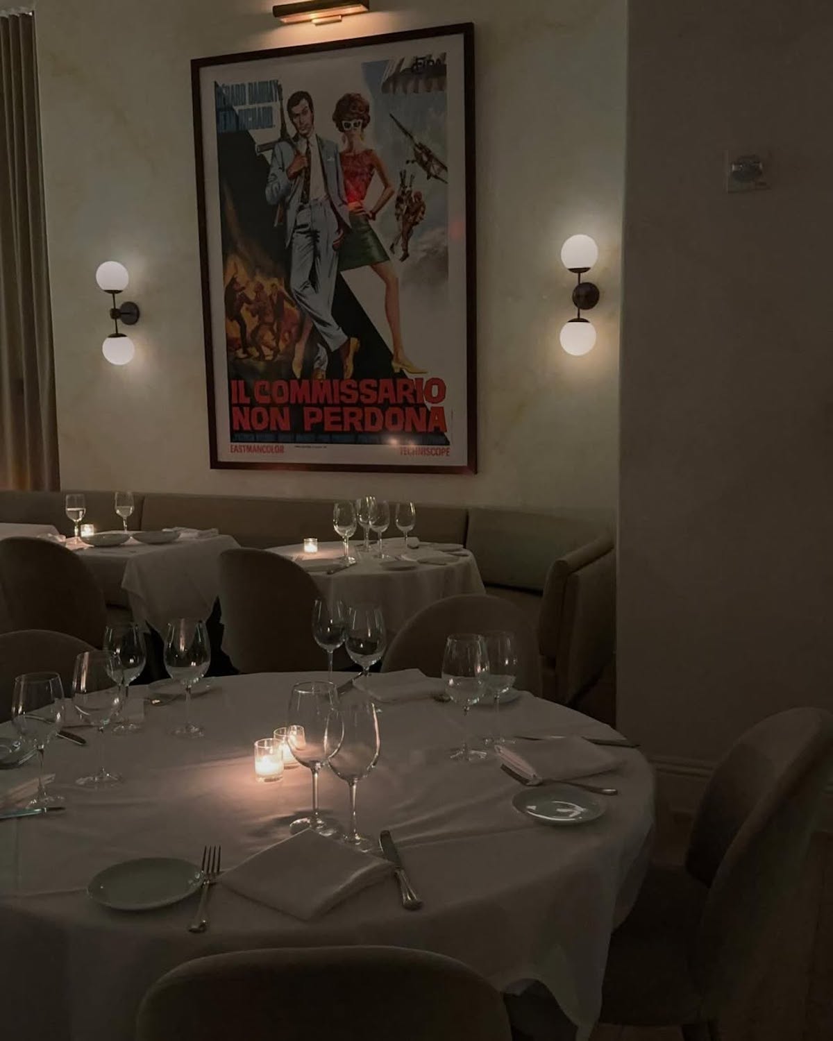 Eleganckie wnętrze restauracji o kameralnym nastroju, oświetlone delikatnym światłem świec. Na pierwszym planie znajduje się stół z białym obrusem, nakryciami, kieliszkami i sztućcami. W tle widoczny jest duży, stylizowany plakat filmowy w języku włoskim, przedstawiający kobietę i mężczyznę z podpisem „Il commissario non perdona”. Pomieszczenie ma przytulny i romantyczny klimat.