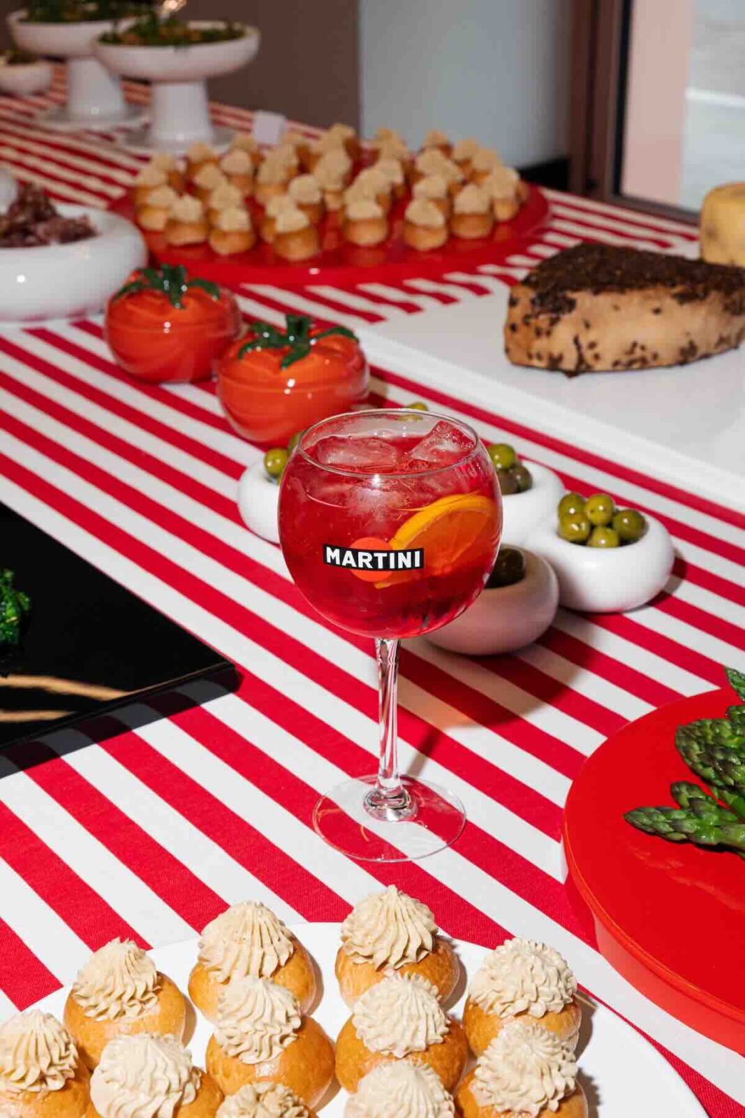 Martini x Mateo Zielonka: Stół pełen włoskich przysmaków: sery, oliwki, przekąski i kieliszek czerwonego drinka Martini Fiero z pomarańczą, wszystko ułożone na charakterystycznym obrusie w biało-czerwone paski.