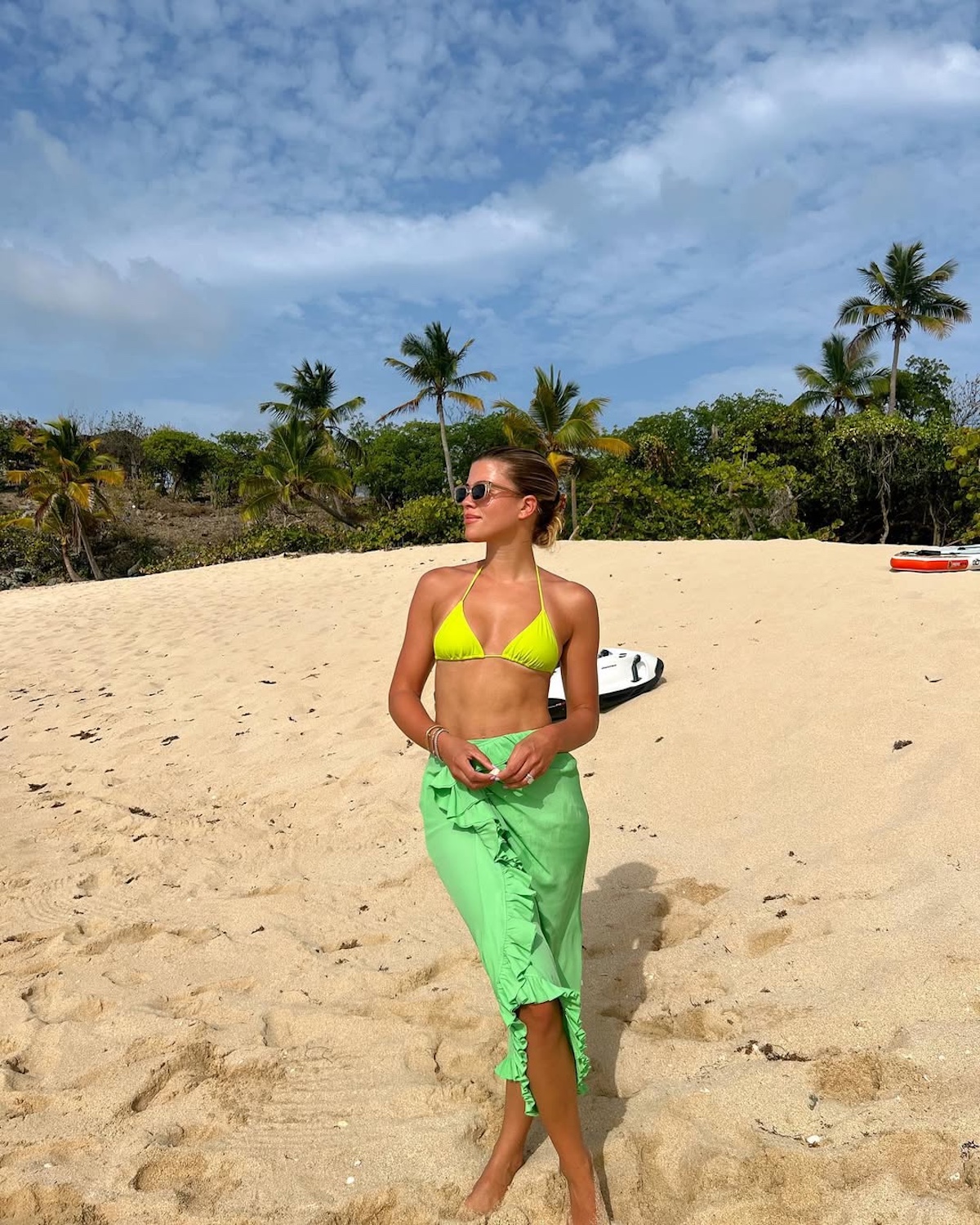 Najlepsze samoopalacze z Rossmanna. Zdjęcie przedstawia kobietę – Sofię Richie Grainge – pozującą na plaży w neonowym bikini i zielonej spódnicy.