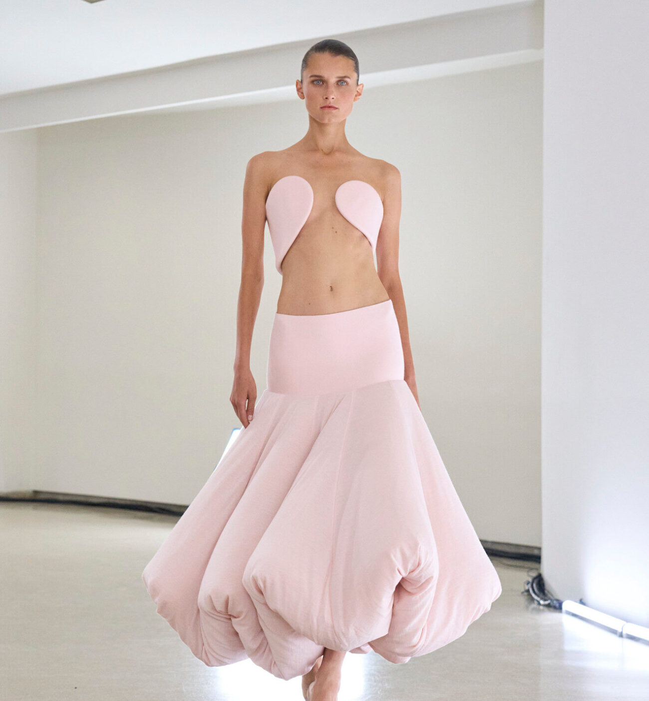 Modelka prezentuje awangardową, jasnoróżową kreację z kolekcji haute couture. Góra stroju składa się z minimalistycznego, rzeźbiarskiego topu o organicznych kształtach, odsłaniającego brzuch. Dół to obszerna, balonowa spódnica o miękkiej strukturze, zakończona zwiniętymi fałdami. Stylizacja jest nowoczesna i artystyczna, modelka idzie boso po jasnej, minimalistycznej przestrzeni.