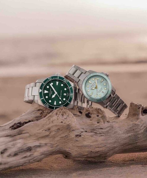CERTINA DS Action: Zielony i turkusowy zegarek Certina DS Action położone na drewnie dryfującym na plaży Zielony i turkusowy model zegarków Certina DS Action z bransoletami ze stali nierdzewnej opierają się o kawałek wyrzuconego na brzeg drewna; kompozycja w naturalnym świetle zachęca do eksplorowania elegancji w stylu outdoor.