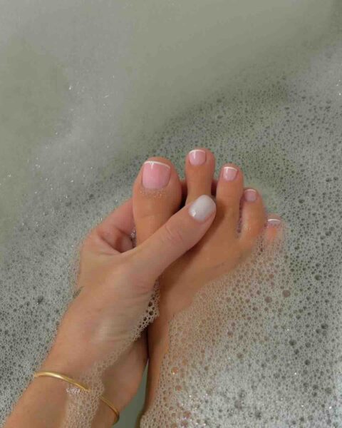 Pedicure na ciepłe dni: Pedicure w stylu francuskim z delikatnym różem i białymi końcówkami, z akcentem białego ombre na jednym palcu, zanurzone w pianie kąpielowej – połączenie relaksu i elegancji.