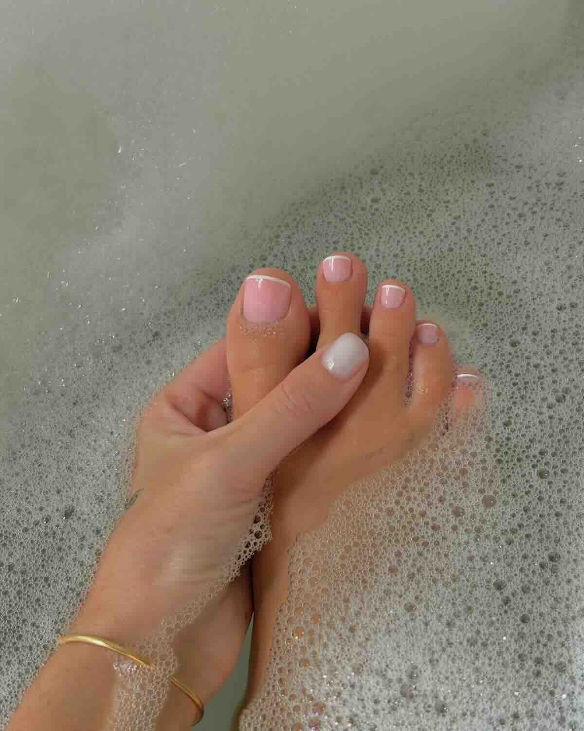 Pedicure na ciepłe dni: Pedicure w stylu francuskim z delikatnym różem i białymi końcówkami, z akcentem białego ombre na jednym palcu, zanurzone w pianie kąpielowej – połączenie relaksu i elegancji.