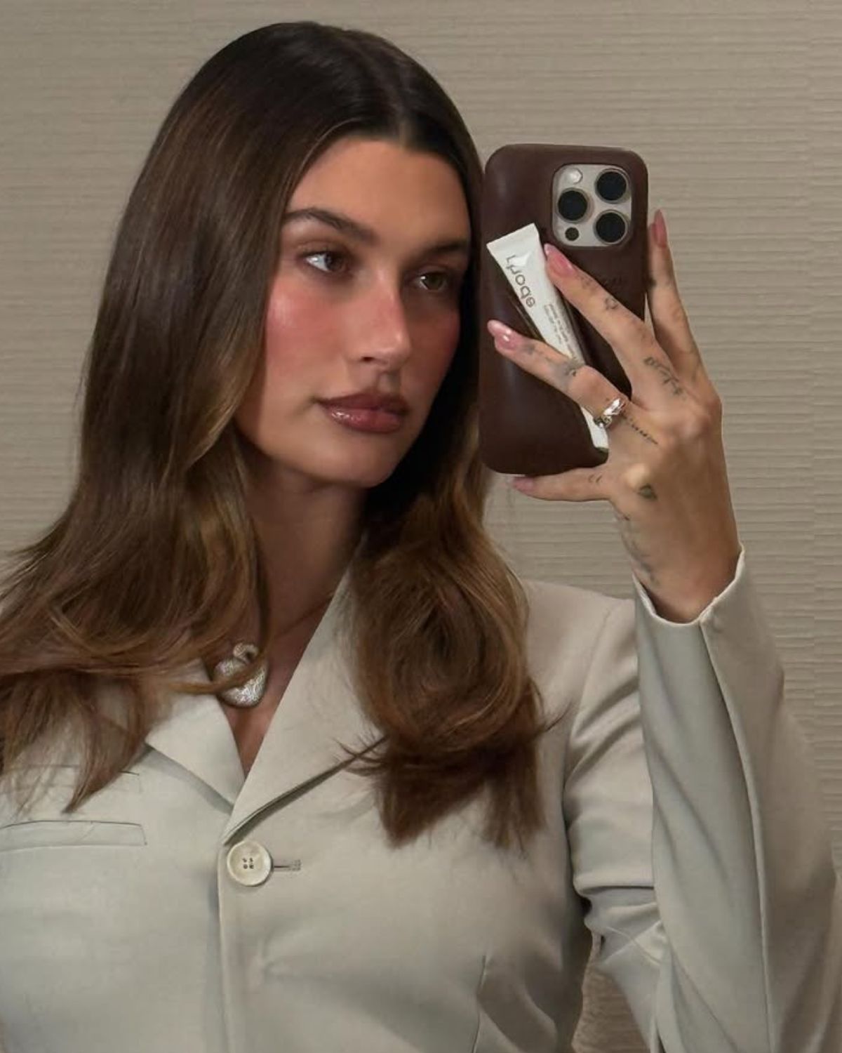 Zdjęcie przedstwiające manicure w stylu Hailey Bieber. Na zdjęciu jest celebrytka ubrana w oliwkową marynarkę. Robi sobie selfie telefonem. Widać jej paznokcie glazed donut. Jej brązowe włosy są rozpuszczone na ramiona.