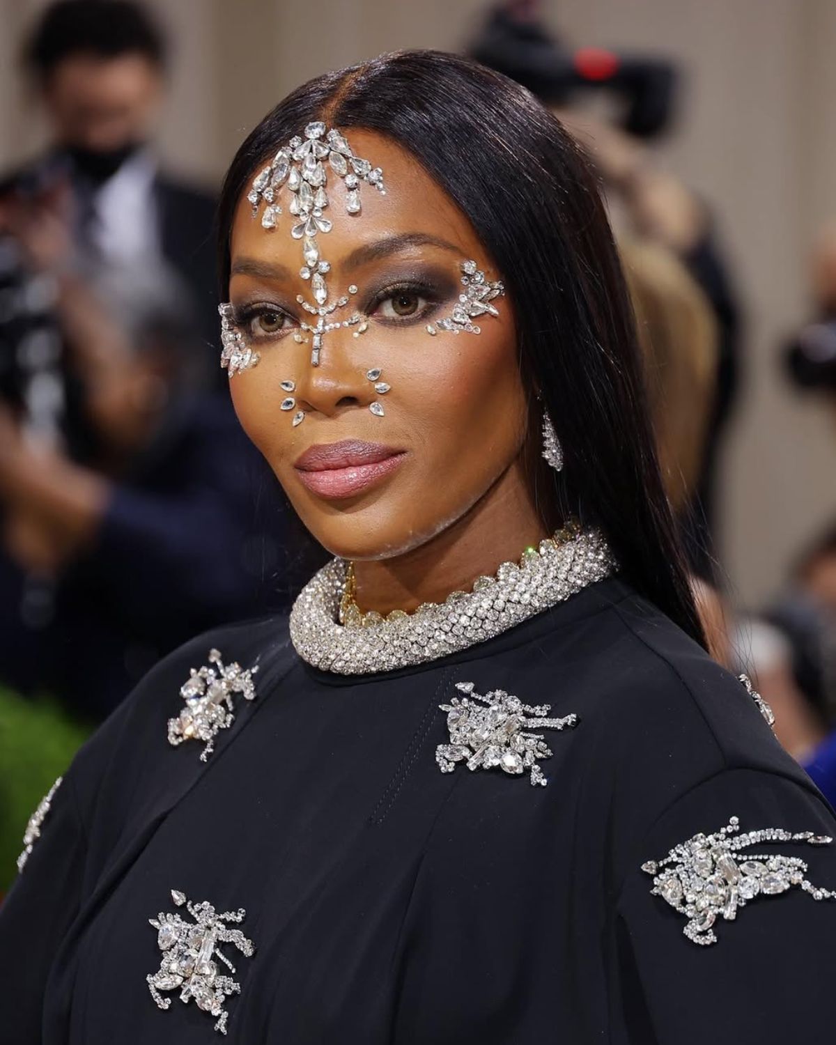 Zdjęcie Naomi Campbell Met Gala 2022. To portret, ze zbliżeniem na jej twarz wysadzaną kamieniami. Ma do tego proste włosy i czarną kreację z masywnym, kryształowym naszyjnikiem.