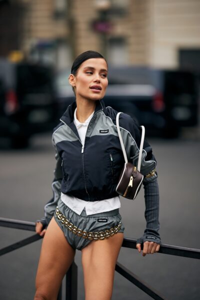 Ubrania na maj 2025: Uliczna stylizacja z Paris Fashion Week: kobieta w sportowym zestawie marki Miu Miu – krótka kurtka, majtki z błyszczącym łańcuchem, białe warstwy pod spodem i miniaturowa torebka przewieszona przez ramię – nowoczesny mix athleisure z luksusem.
