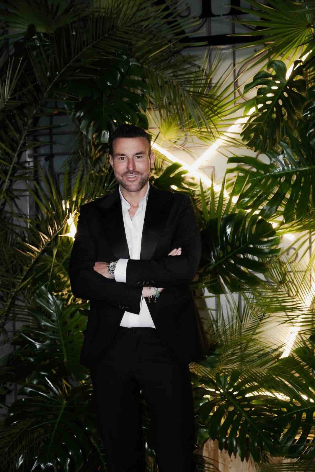 Philipp Plein nowy butik: Mężczyzna w eleganckim czarnym garniturze stoi na tle bujnych tropikalnych liści z neonowym oświetleniem, z uśmiechem i rękami skrzyżowanymi na piersi, gotowy na wieczorne wydarzenie modowe.