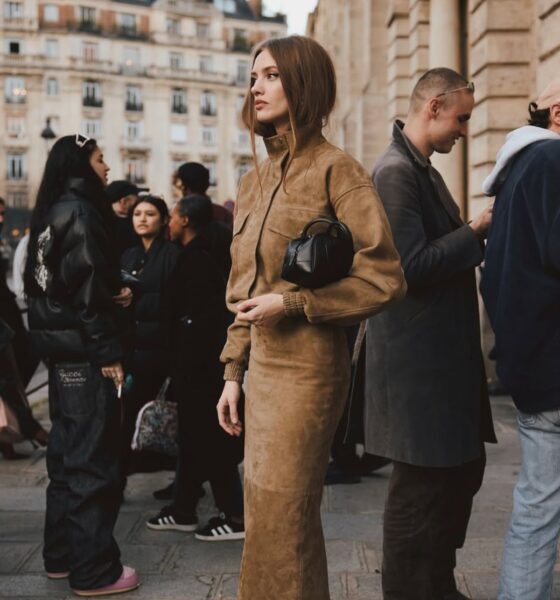 Na zdjęciu znajduje się Sylwia Butor @sylwia_butor. Ubrana jest w zamszowy komplet z kurtką i spódnicą maxi w kolorze beżowym. Na nogach ma czarne sandałki. Pod ręką czarna minitorebka. Stoi na ulicy w tłumie.