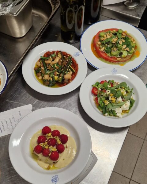 Widok z góry na blat kuchenny, na którym znajdują się cztery estetycznie podane potrawy na białych talerzach. Potrawy to kolorowe, apetycznie wyglądające dania, w tym deser z malinami i pistacjami, świeże sałatki z warzywami i orzechami oraz grillowane warzywa. Obok talerzy widoczne są butelki oraz karteczka z odręcznie zapisanymi notatkami. Całość sprawia wrażenie świeżej, sezonowej kuchni.