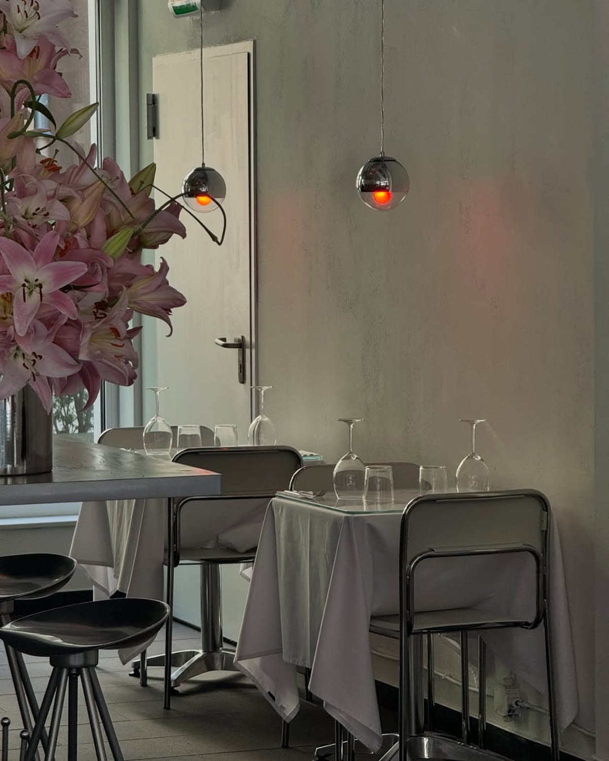 Minimalistyczne wnętrze restauracji z metalowymi krzesłami i stołami przykrytymi białymi obrusami. Na stołach ustawione są odwrócone kieliszki i szklanki. Po lewej stronie widoczny bukiet różowych lilii w metalowym wazonie.