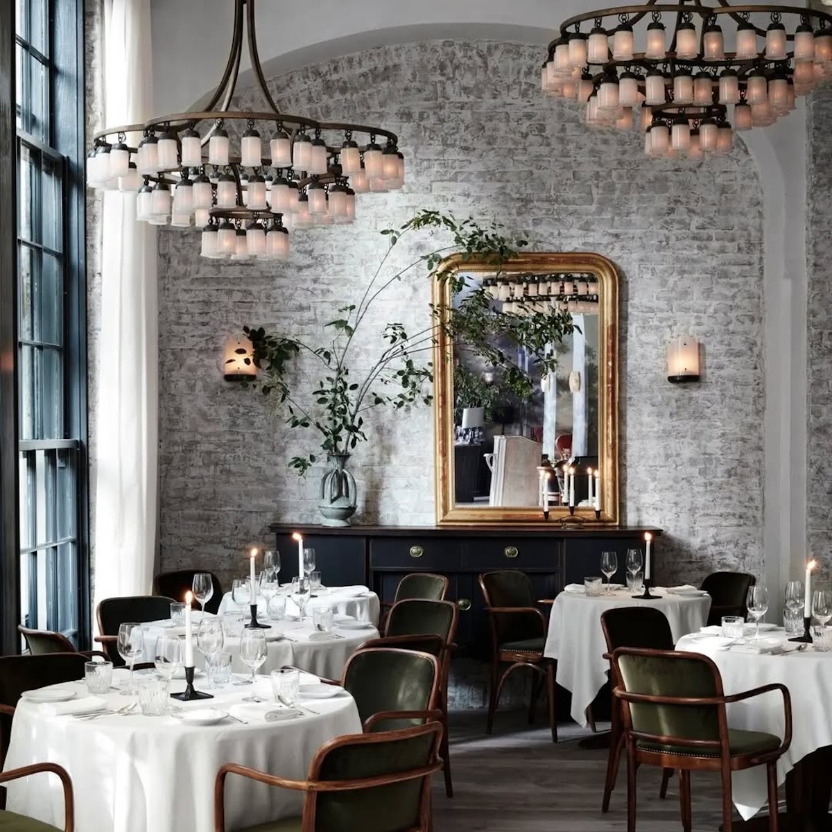 Restauracje otwarte w święta wielkanocne 2025. eleganckie wnętrze restauracji z elementami stylu vintage. Na pierwszym planie znajdują się okrągłe stoliki przykryte białymi obrusami, wokół których ustawione są ciemnozielone, welurowe krzesła. Na każdym stole stoi świecznik ze świecą, kieliszki i zastawa. W tle widoczna jest ściana z jasnej, nieregularnej cegły, na której zawieszone jest duże, złote lustro w ozdobnej ramie oraz ciemna komoda z wazonem i gałązkami.